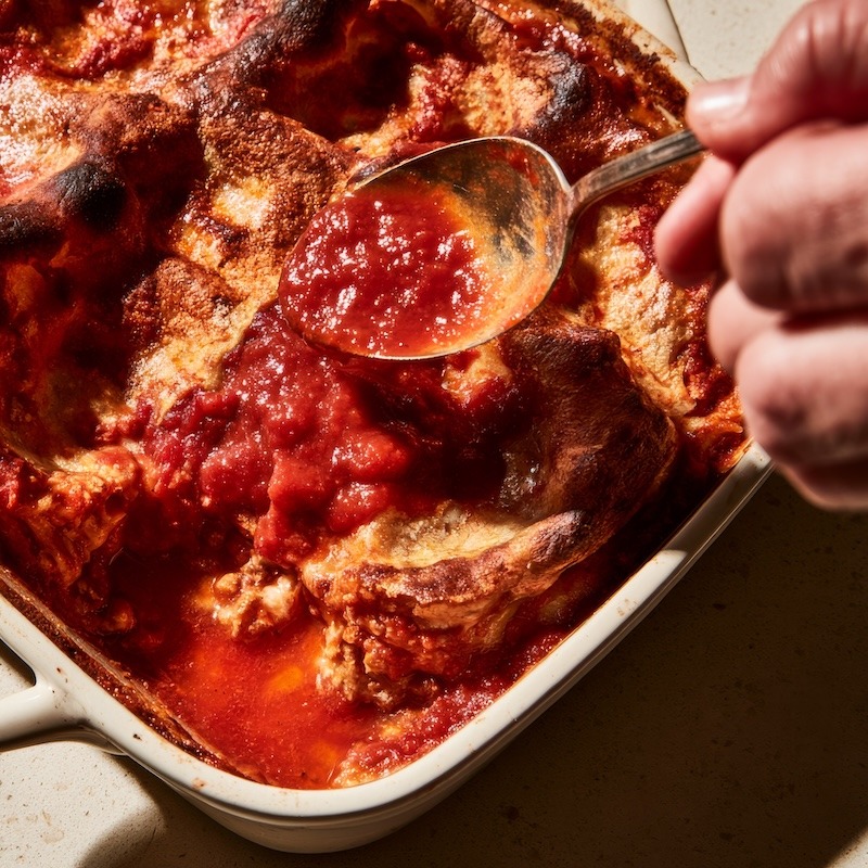 A spoon drizzling extra tomato sauce over the dry, hardened top layer of a baked lasagna