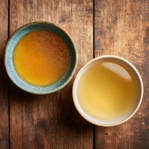 Two bowls side by side on a rustic wooden countertop, left bowl containing golden, rich chicken stock, right bowl containing light, clear chicken broth.