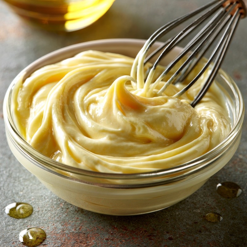 A small bowl of finished, glossy pale-yellow mayonnaise with a whisk resting against the side