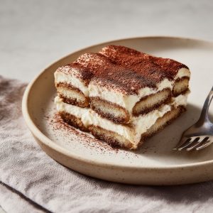 A cleanly sliced tiramisu in a shallow ceramic dish reveals airy mascarpone cream layered with lightly coffee-dipped ladyfingers, finished with a fresh dusting of cocoa