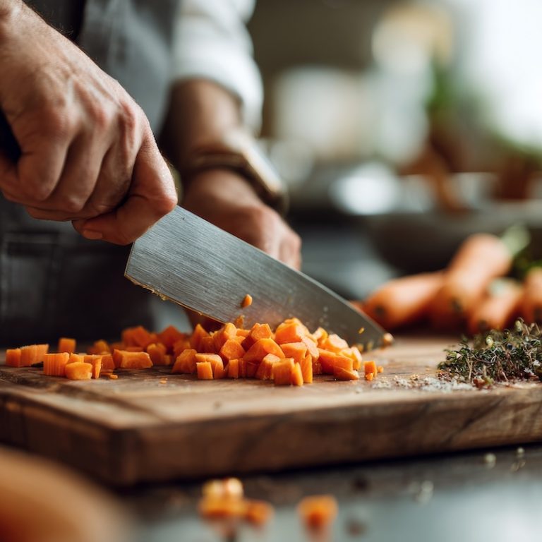A stainless steel chefs knife, dicing carrots on a wooden cutting board.