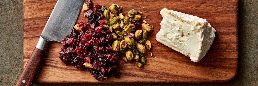 wooden cutting board with roughly chopped dried cherries, toasted pistachios, and a wedge of fresh goat cheese