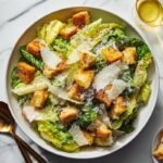 Classic Caesar salad plated in a wide shallow white bowl, whole romaine leaves rather than chopped, shaved Parmesan curls