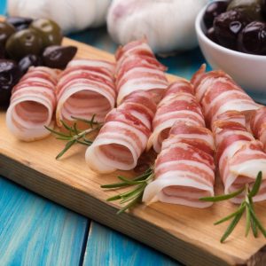 A wooden cutting board with rolled slices of pancetta and rosemary.
