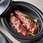 a slow cooker insert with two raw pork tenderloins nestled in broth with garlic and rosemary sprigs
