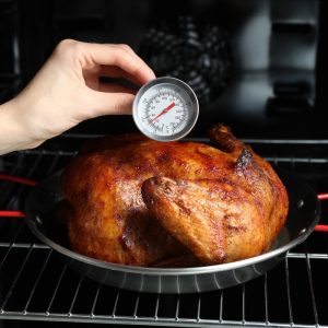 Whole chicken roasting in the oven with an analog thermometer measuring internal temperature.