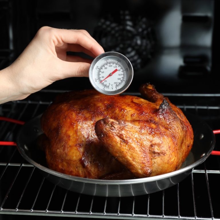 Whole chicken roasting in the oven with an analog thermometer measuring internal temperature.