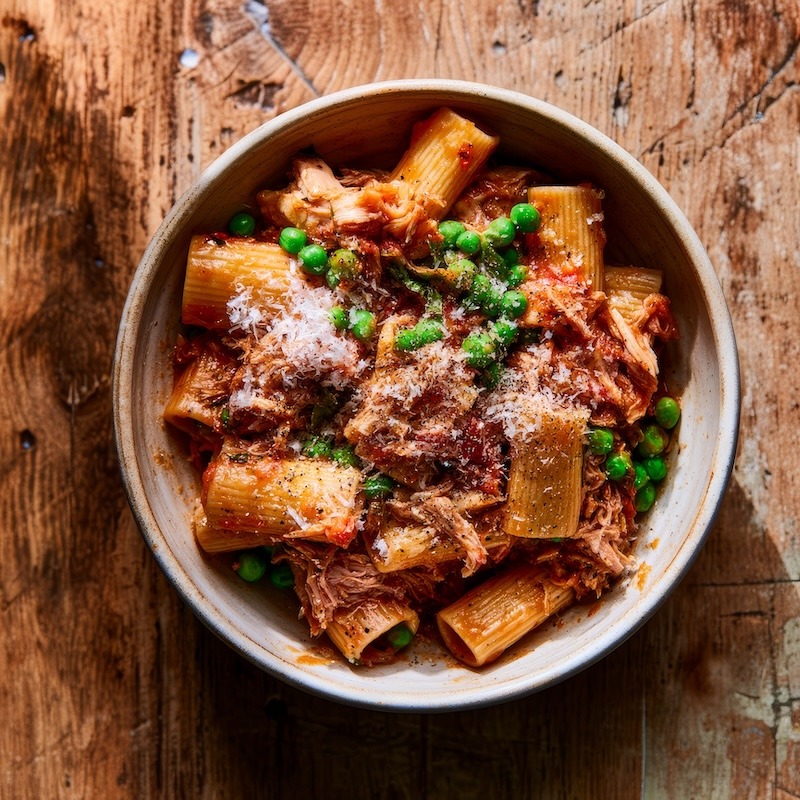 A bowl of rigatoni with shredded rotisserie chicken, bright green peas, and a glossy tomato basil sauce