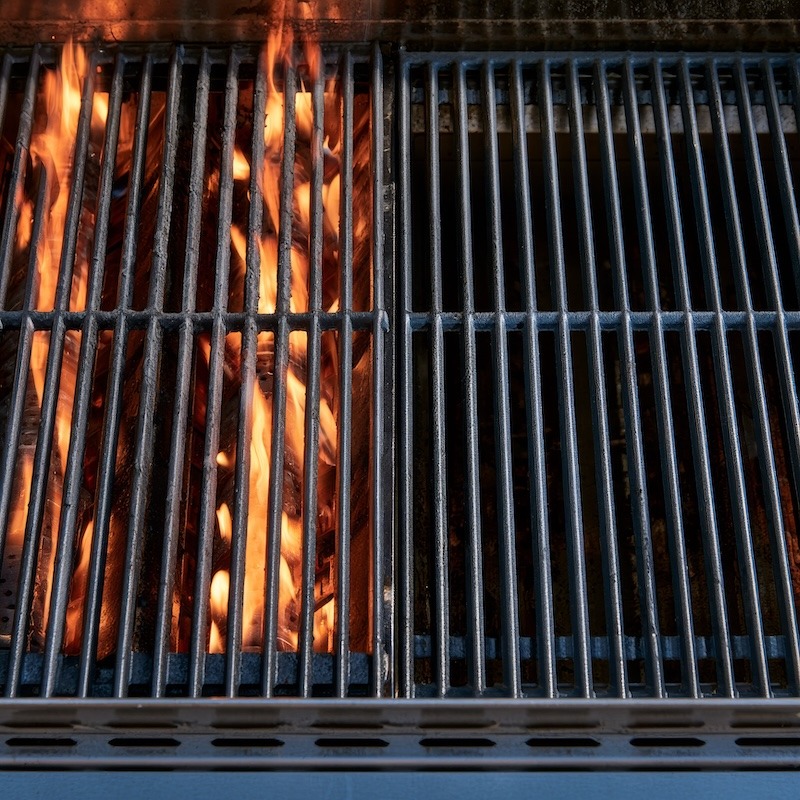 a gas grill with visible flame under the burners on one side only, the grates on the other side clean and empty with no flame beneath them
