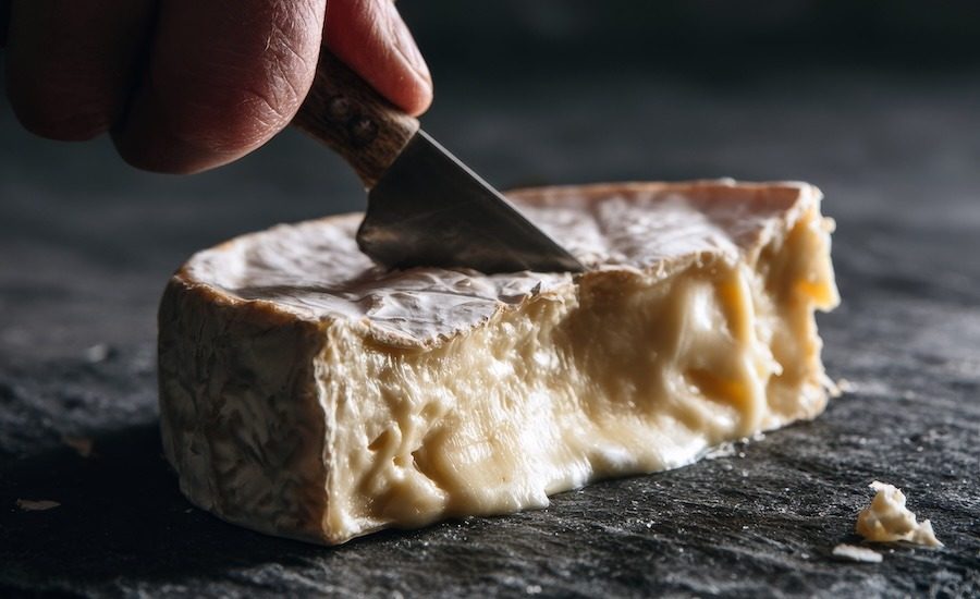 a hand holding a small knife cutting through a wedge of ripe Brie