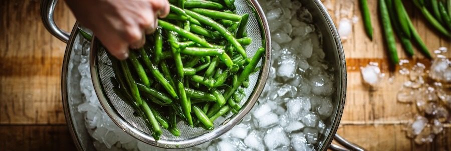 plunging a metal strainer of bright green green beans into a large bowl of ice water